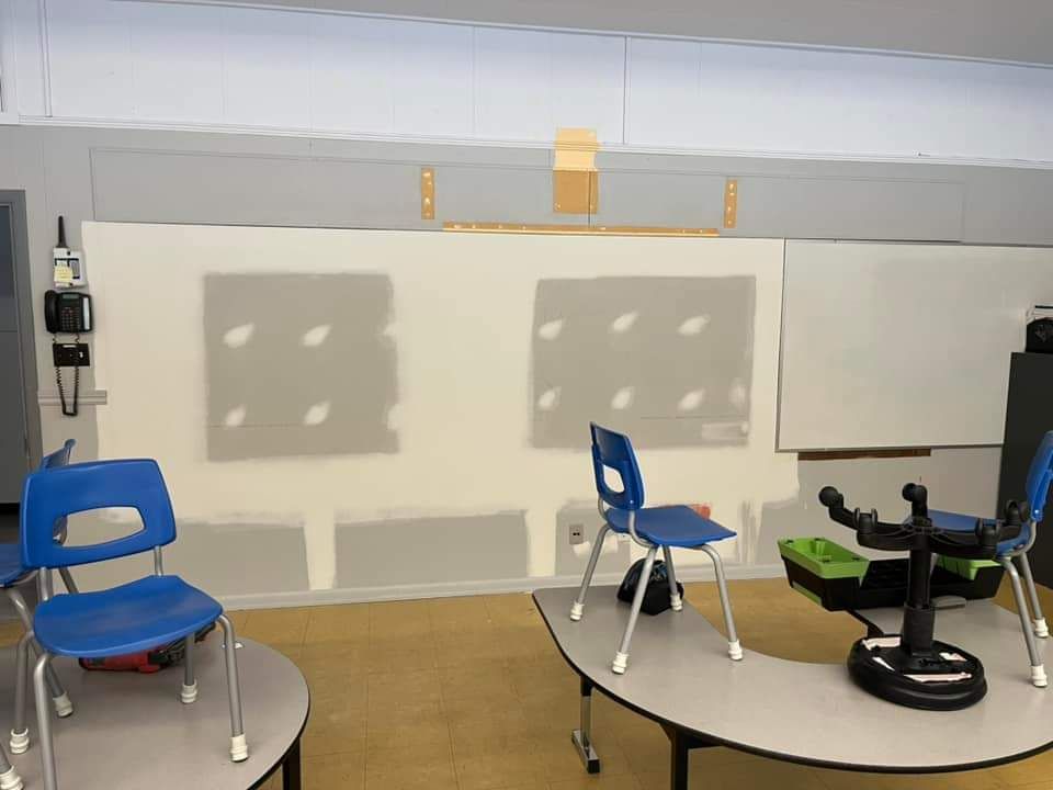Salle de classe avec chaises bleues, tables, tableau blanc et mur partiellement fini.