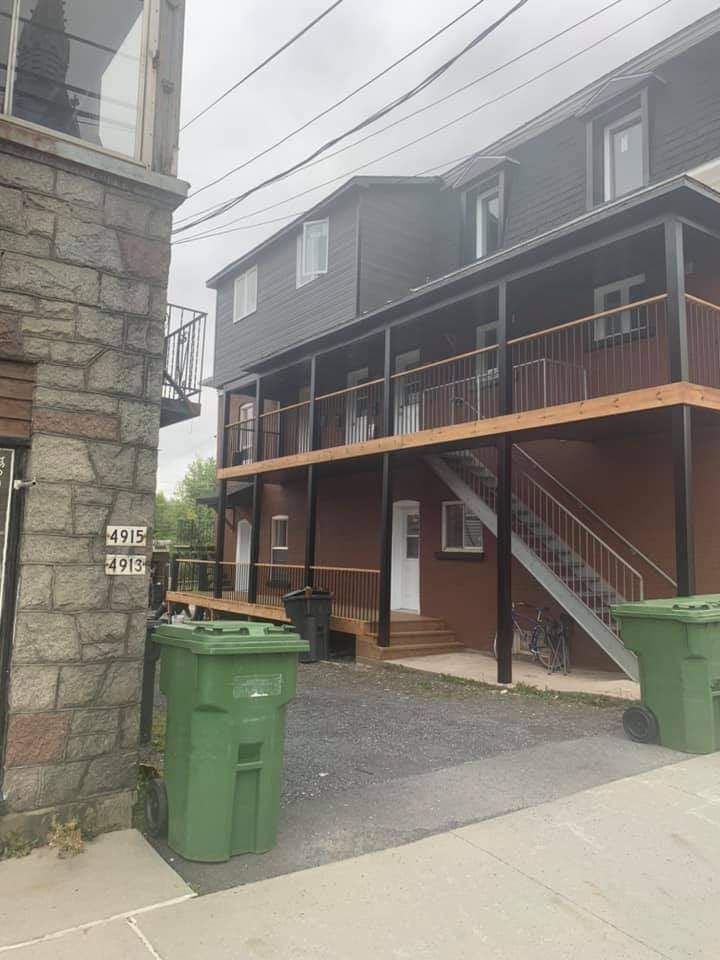 Immeuble à plusieurs étages avec escaliers extérieurs et balcons, jouxtant une structure en pierre avec des poubelles vertes au premier plan.