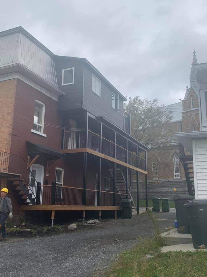 Extérieur d'un bâtiment avec un bardage foncé et des terrasses en bois. Une personne portant un casque de chantier se tient à proximité.