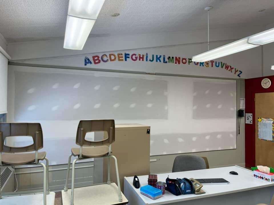 Salle de classe avec les lettres de l'alphabet au-dessus d'un tableau blanc, deux chaises et un bureau pour le professeur.