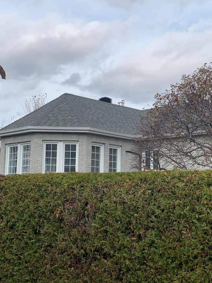 Maison en briques grises avec un toit gris foncé, dissimulée derrière une haie verte par temps couvert.