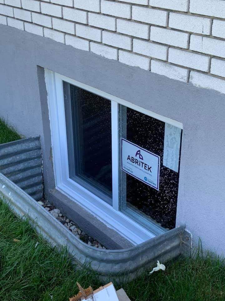 Fenêtre de sous-sol à cadre blanc dans un mur gris, installée avec un puits de lumière dans la pelouse.