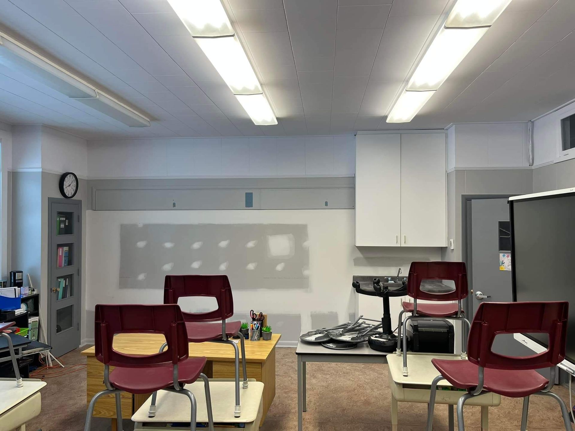 Salle de classe vide avec des chaises rouges, un tableau blanc et des armoires. Des néons éclairent la pièce.