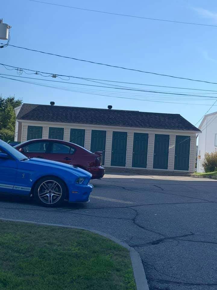 Une Ford Mustang bleue est garée dans la rue, devant un bâtiment blanc aux volets verts et une voiture rouge.