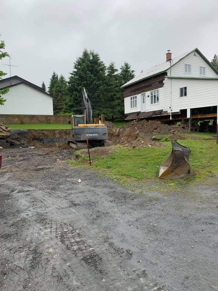 Une excavatrice creuse près d'une maison à deux étages, probablement pour des travaux de construction, par temps nuageux.