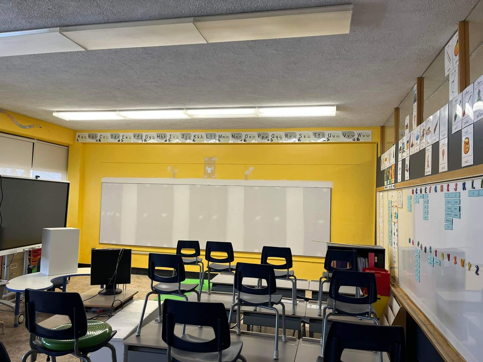 Salle de classe vide avec des murs jaunes, des tableaux blancs et des chaises noires.