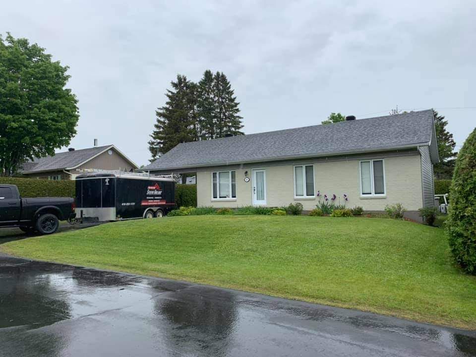 Maison de style ranch avec toit gris, pelouse verte et caravane noire par temps de pluie.