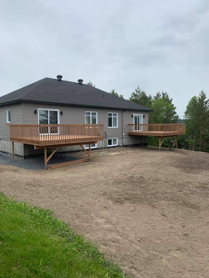 Maison avec deux terrasses en bois ; bardage gris clair, toit foncé et cour en terre battue.