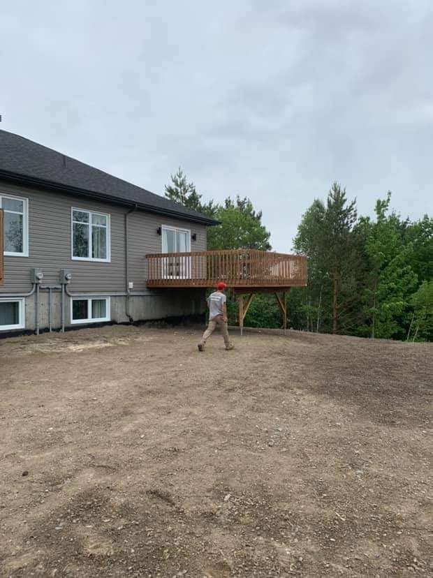 Personne s'éloignant d'une maison avec une terrasse en bois ; journée nuageuse, sol en terre brune.