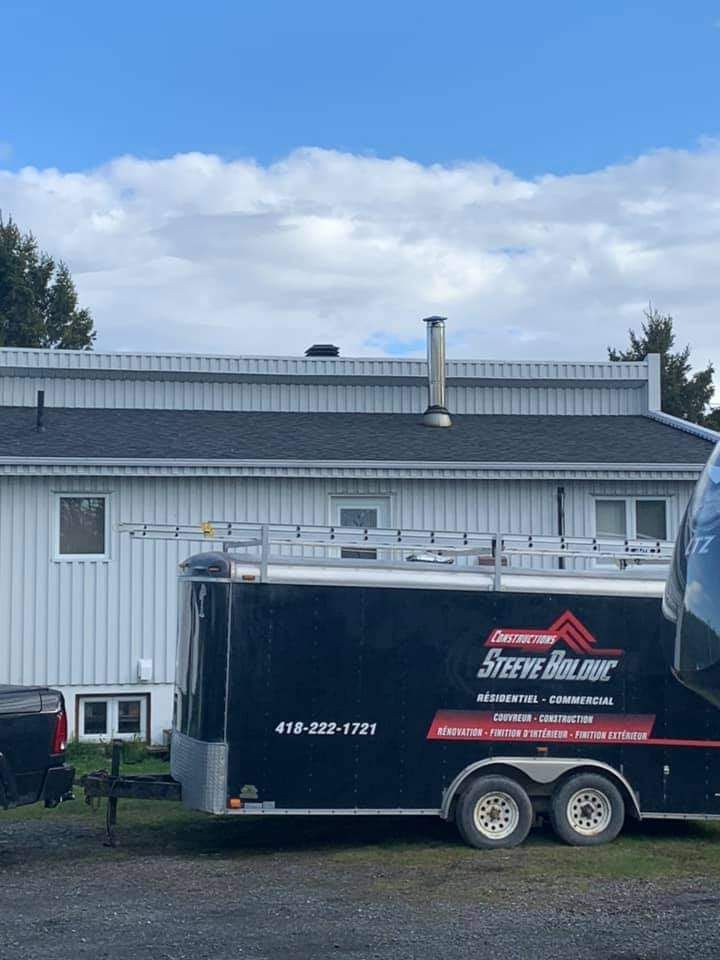 Une remorque noire portant le logo « Steve Builder » est garée près d'une maison dont le toit est neuf.