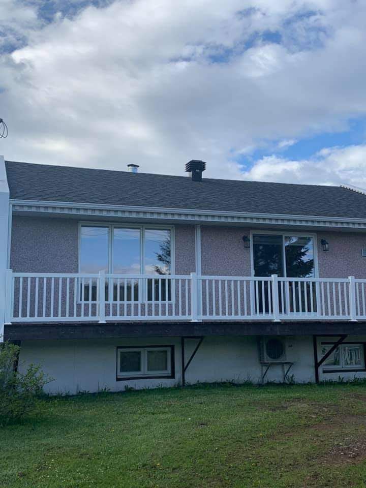 Arrière de la maison avec terrasse blanche, murs roses, toit sombre, deux fenêtres et cheminée sombre sur fond de ciel nuageux.