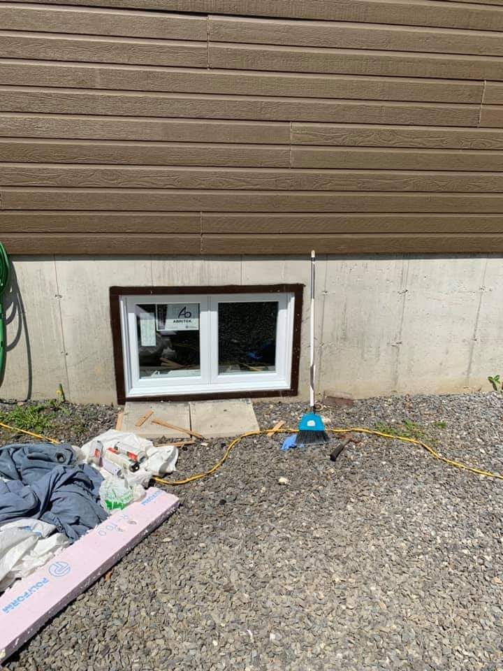 Fenêtre de sous-sol à cadre brun, encastrée dans une fondation en béton sous un revêtement extérieur brun. Sol gravillonné avec débris, balai et tuyau d'arrosage.