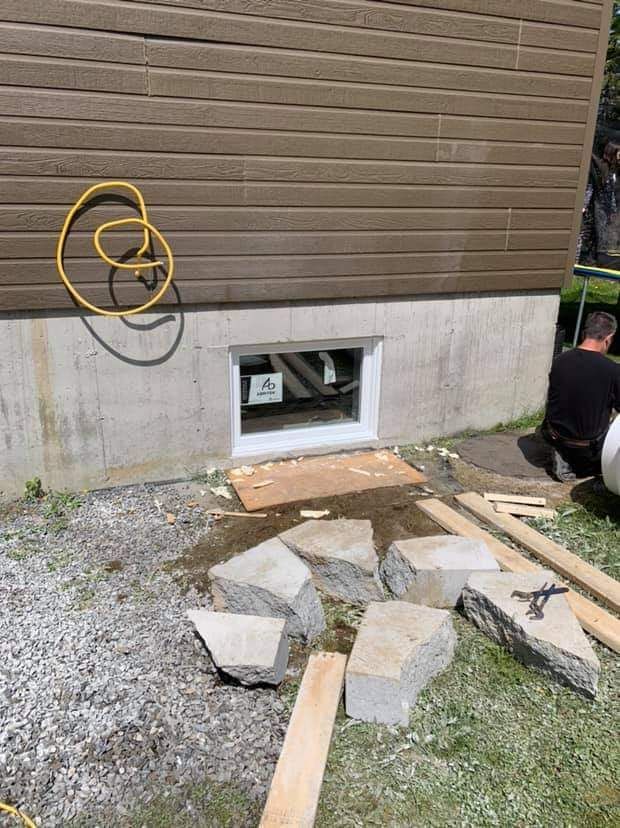 Fondation en béton avec fenêtre. Une personne travaille à l'extérieur, près de pierres et de bois.