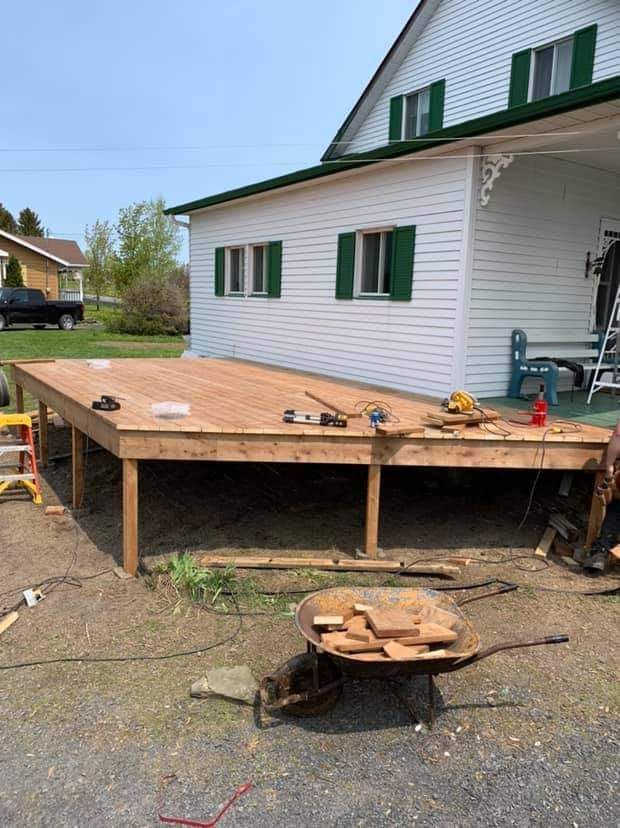 Terrasse en bois neuve attenante à une maison blanche aux volets verts ; une personne travaille à proximité avec des outils et une brouette.