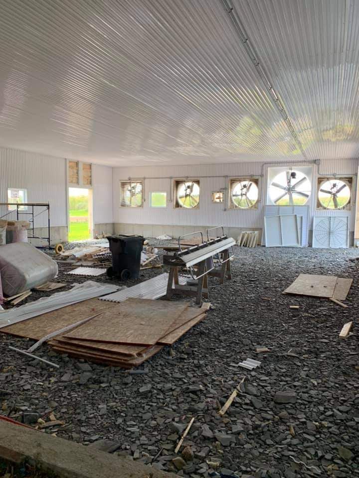 Intérieur d'un grand bâtiment vide avec toit en métal, sol en terre battue, conduits d'aération et matériaux de construction éparpillés.