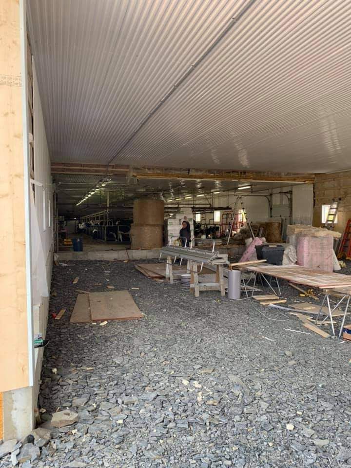 Intérieur d'un bâtiment en construction : sol en gravier, plafond en tôle ondulée. On aperçoit des tables, des outils et une personne.