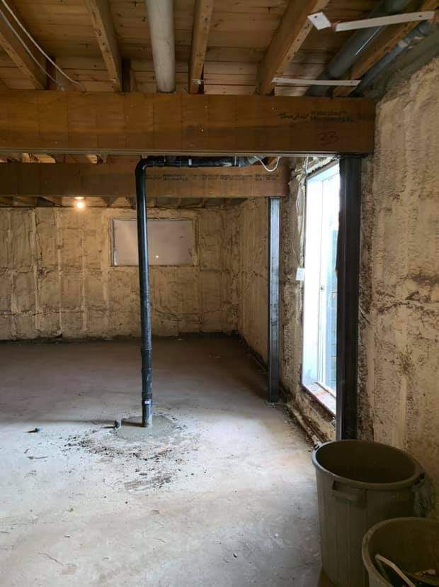 Sous-sol avec poutres apparentes, sol en béton, tuyau noir et porte. Des seaux sont posés à proximité.