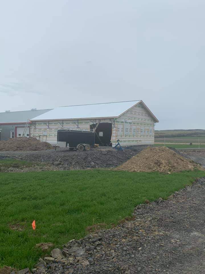 Chantier avec un bâtiment en construction ; un camion est stationné à proximité. Au premier plan, on aperçoit de l’herbe verte et des tas de terre. Ciel couvert.