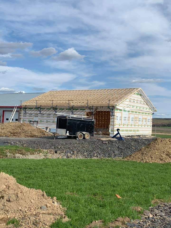 Chantier de construction, bâtiment inachevé avec charpente de toit, entouré de gravier et d'herbe, sous un ciel nuageux.