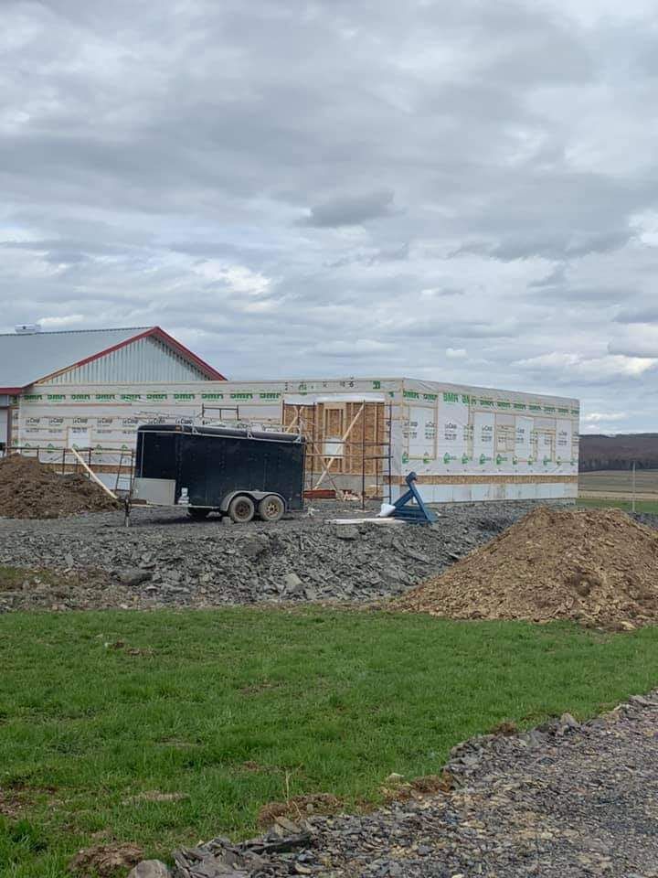 Chantier : bâtiment à ossature bois, bardage blanc. Remorque et tas de terre au premier plan. Ciel couvert.