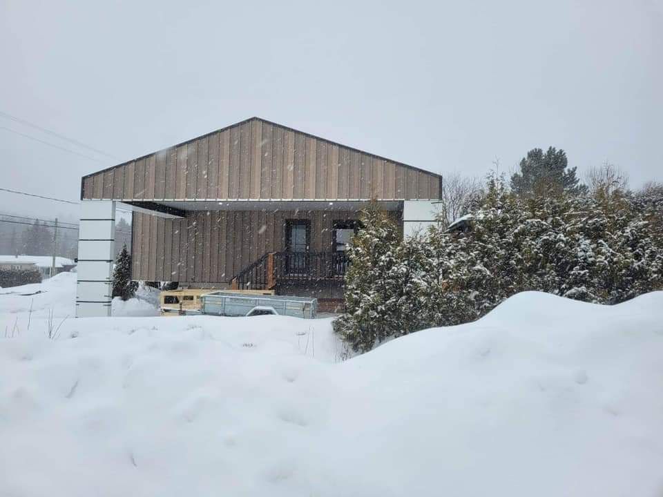 Scène enneigée d'une petite maison à bardage en bois et porche sous une neige qui tombe.
