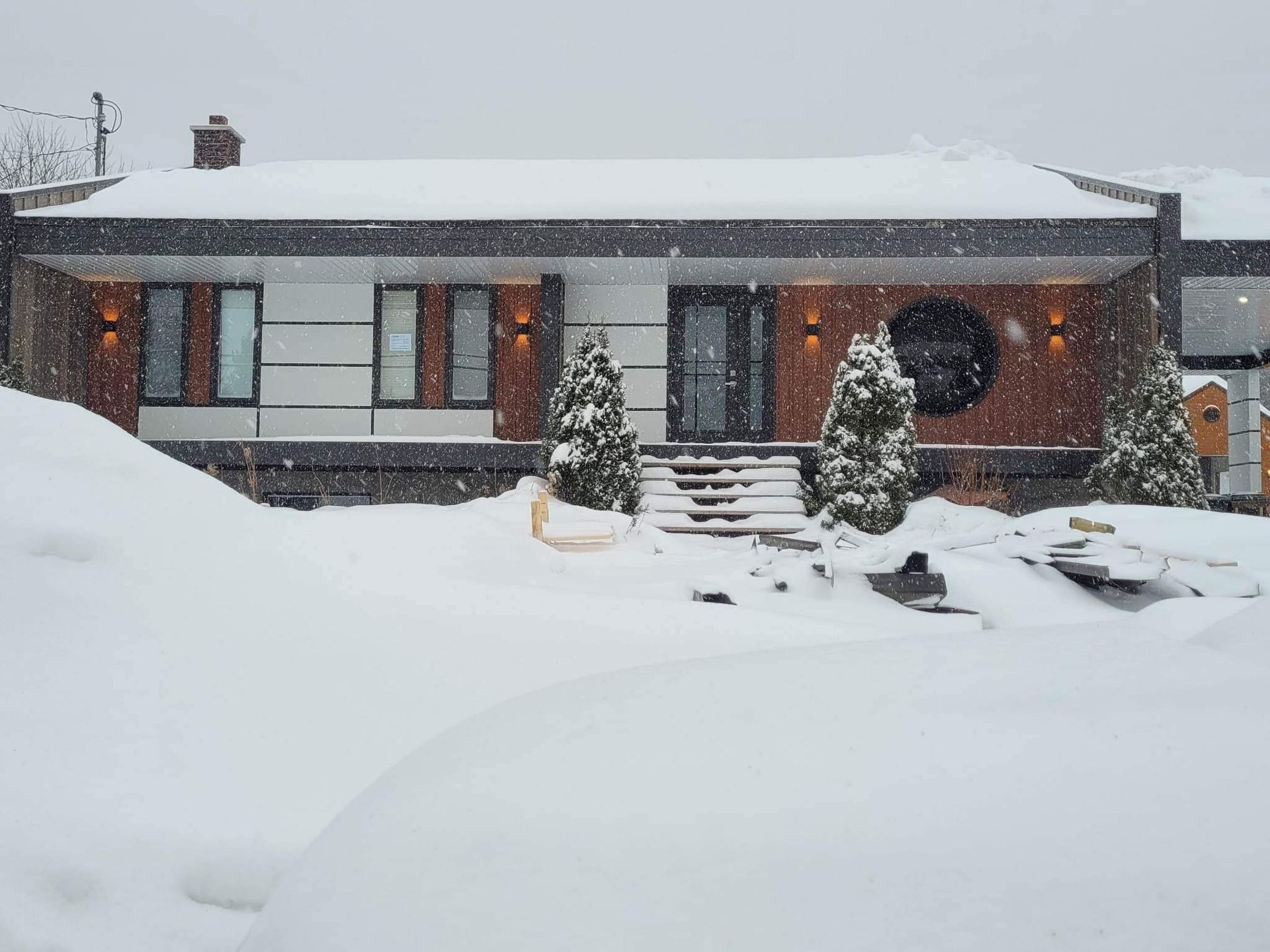 Maison enneigée avec porche couvert, marches et conifères. Une épaisse couche de neige recouvre la cour.