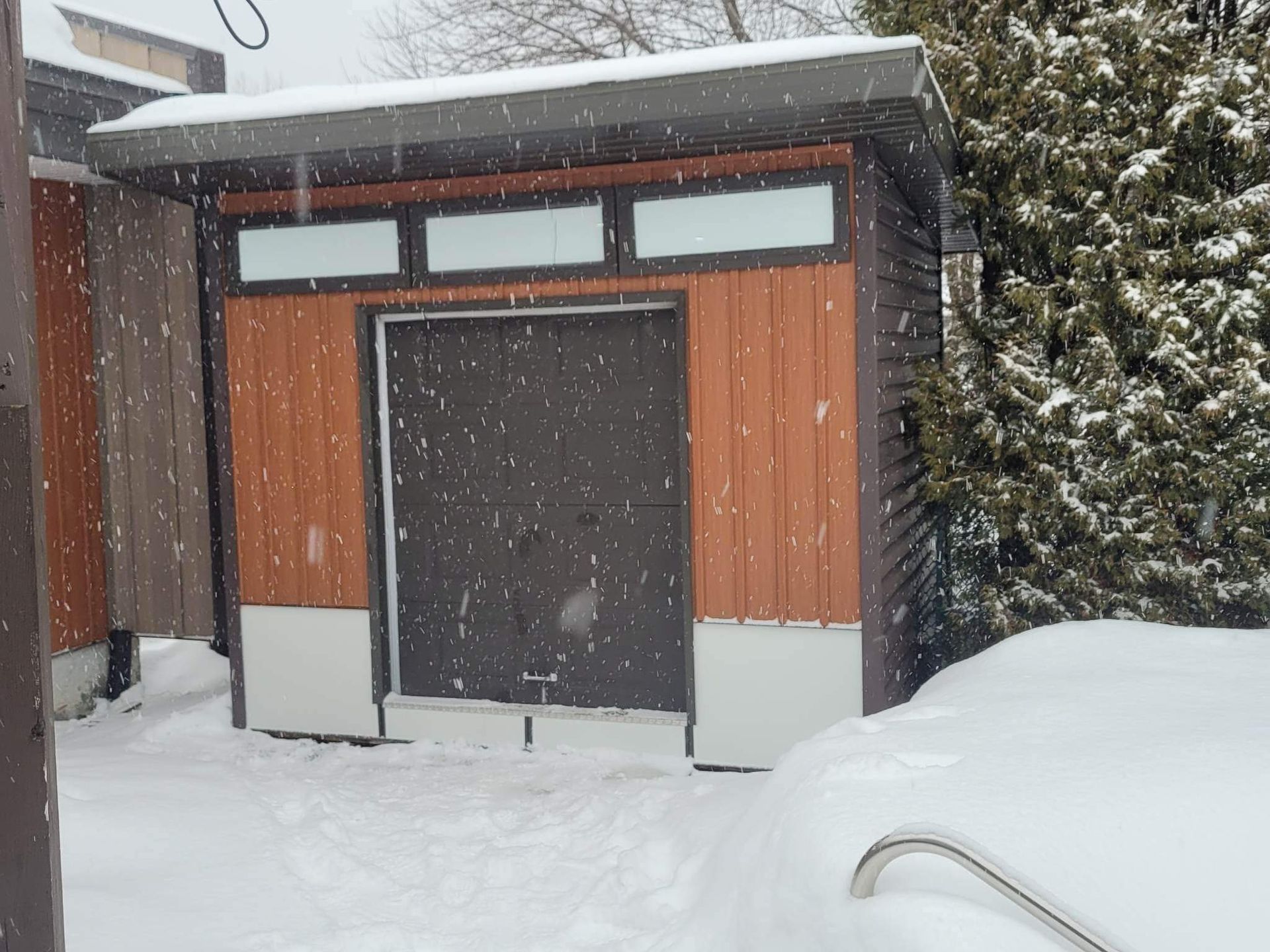 Petit abri de jardin en bois, entouré de neige. Il neige.