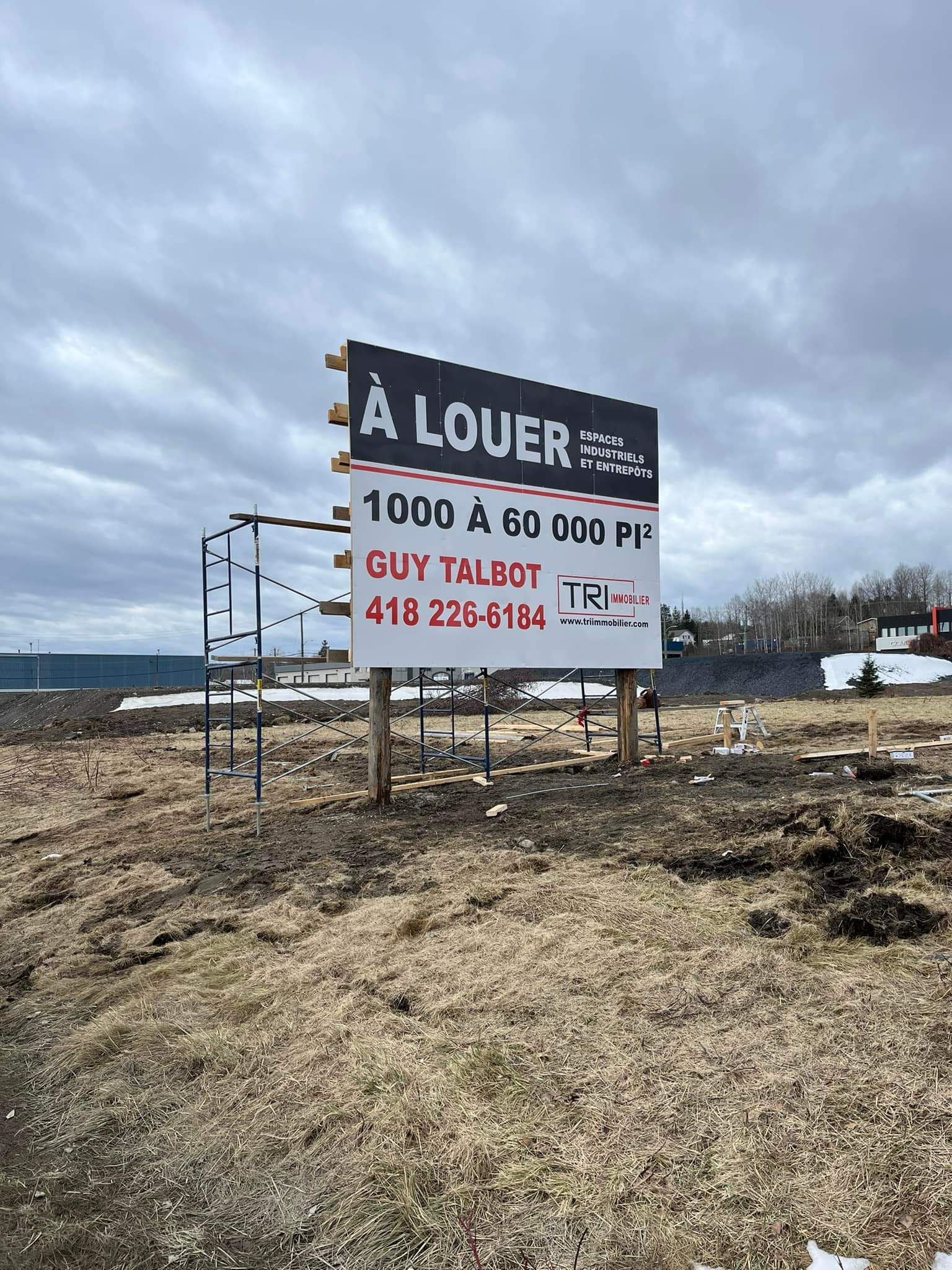 Panneau « À LOUER » (À louer) sur un terrain vacant, avec numéro de téléphone et superficie. Ciel couvert.