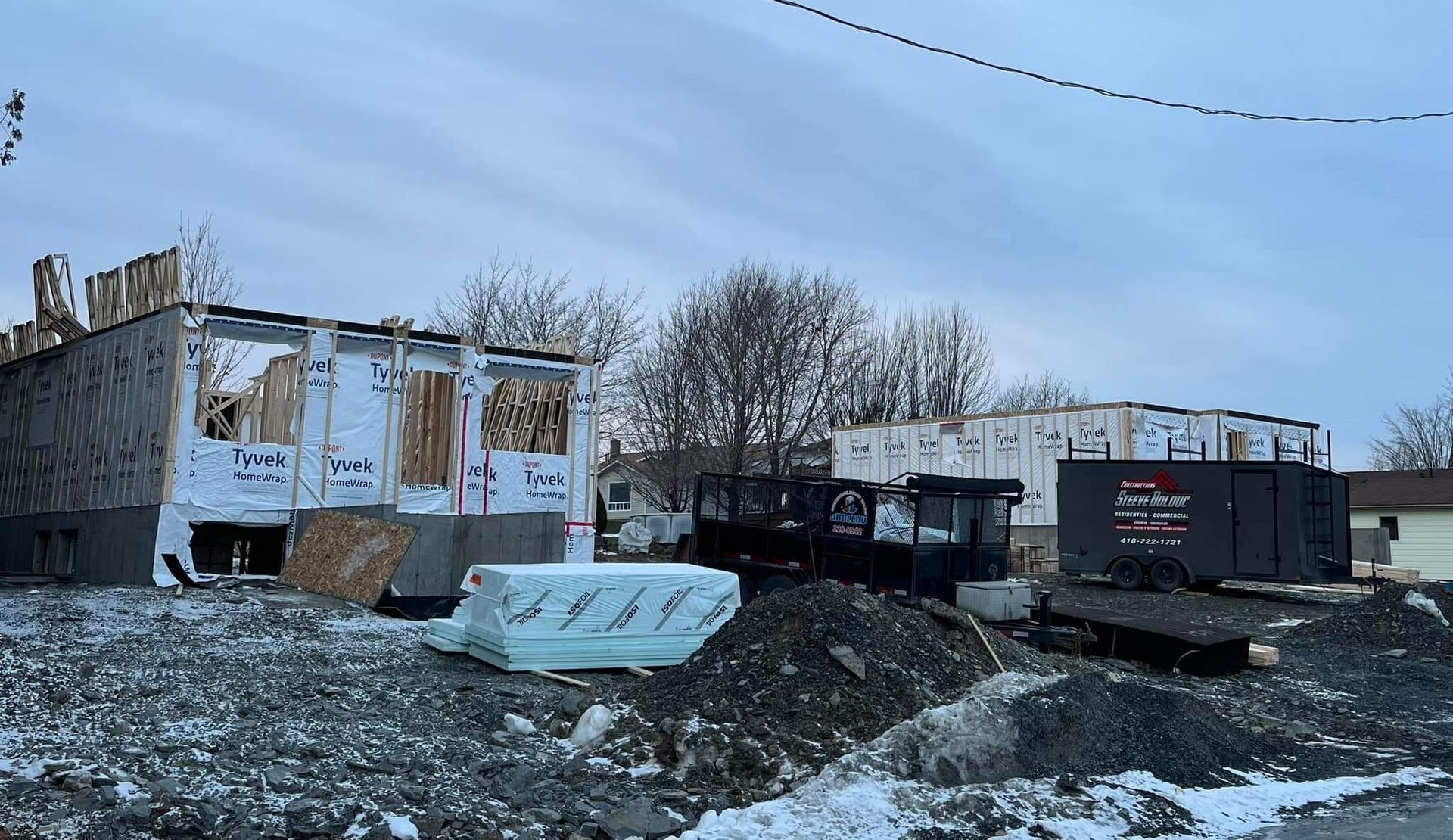 Chantier : structures partiellement construites avec des ossatures en bois, débris et conteneurs. Ciel nuageux.
