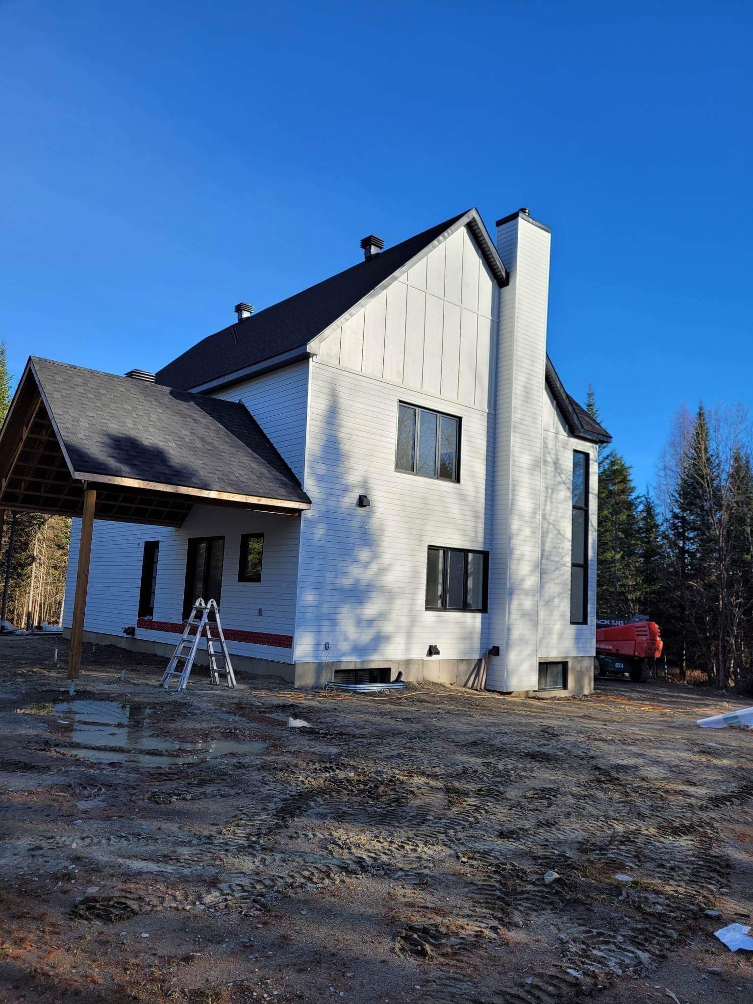 Maison en construction avec bardage blanc, toit noir et cheminée. Journée ensoleillée, entourée d'arbres.