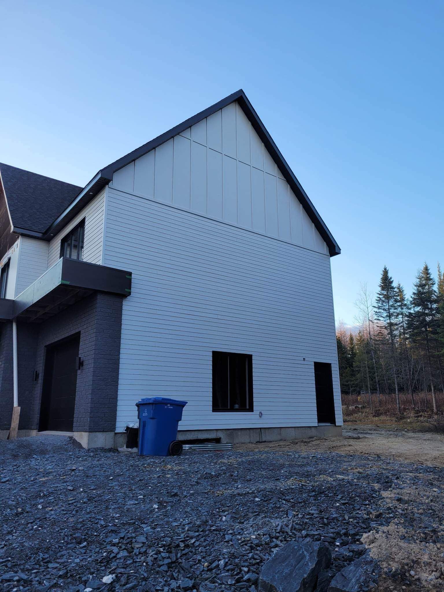 Vue extérieure d'une maison en construction avec un bardage clair, un toit noir et une poubelle bleue.
