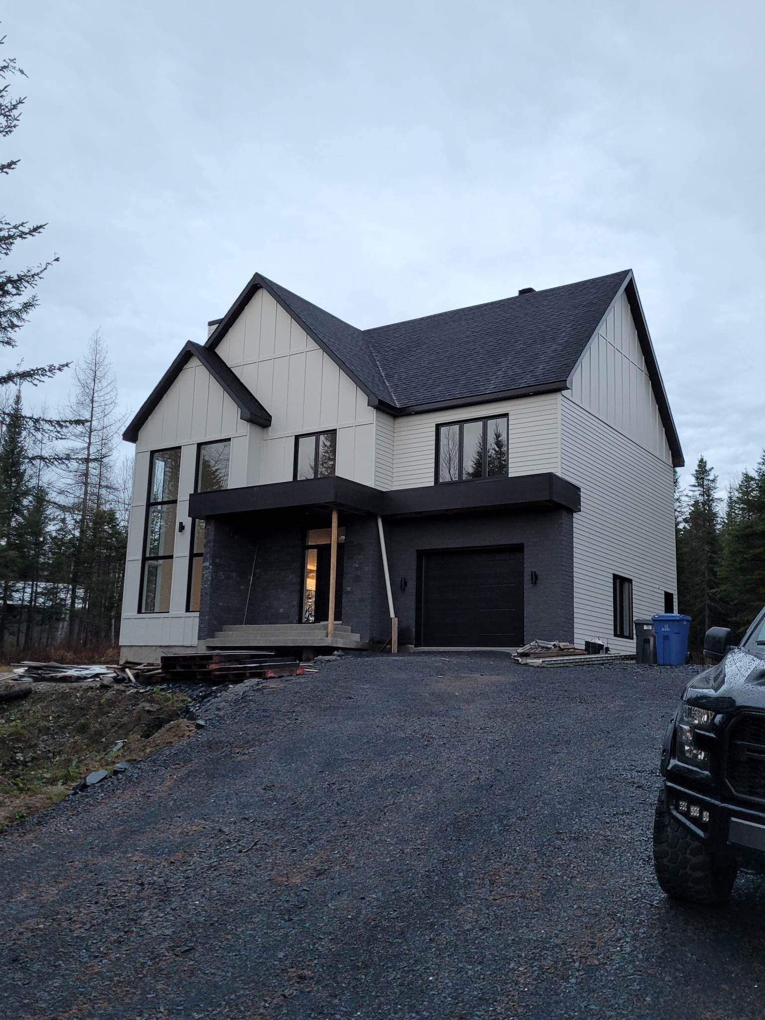 Maison moderne à deux étages avec boiseries et toiture noires. Bardage gris, allée en gravier, ciel couvert.