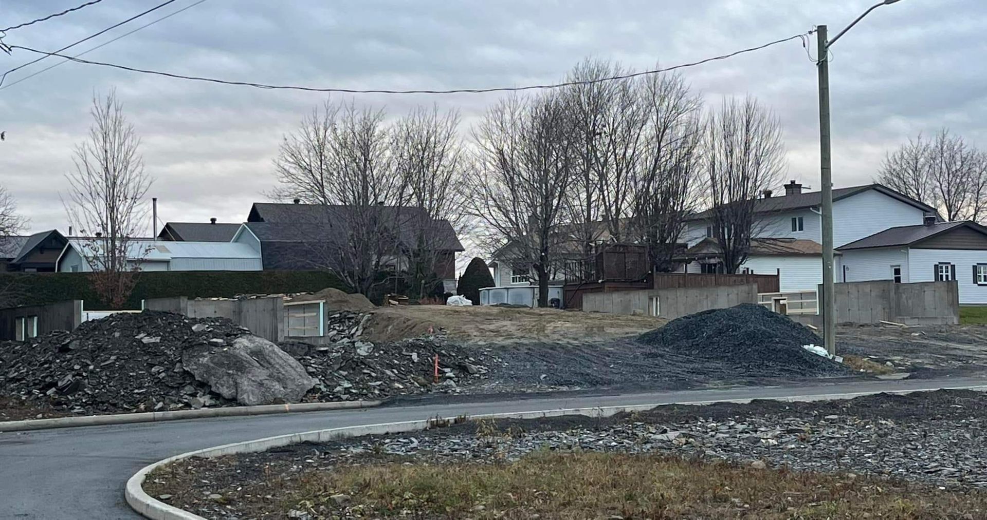 Scène de rue résidentielle avec des tas de gravier, des maisons et des arbres dénudés sous un ciel nuageux.