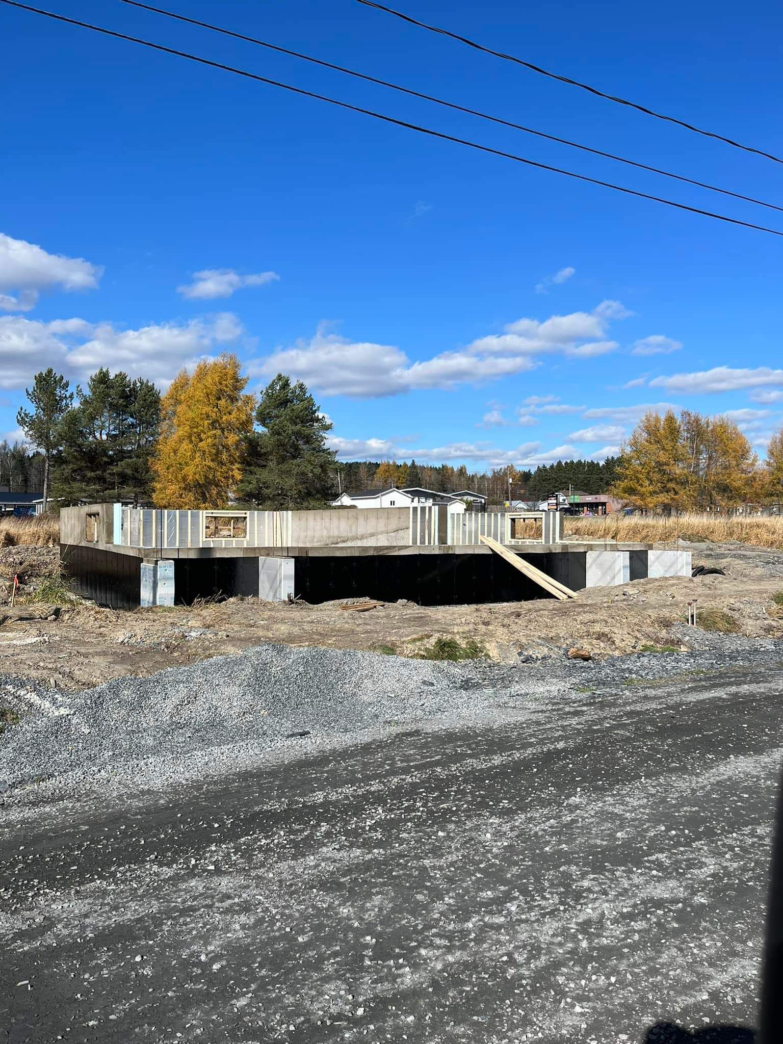 Fondations d'un bâtiment en construction sur un terrain gravillonné ; arbres aux feuilles jaunes en arrière-plan, sous un ciel bleu.