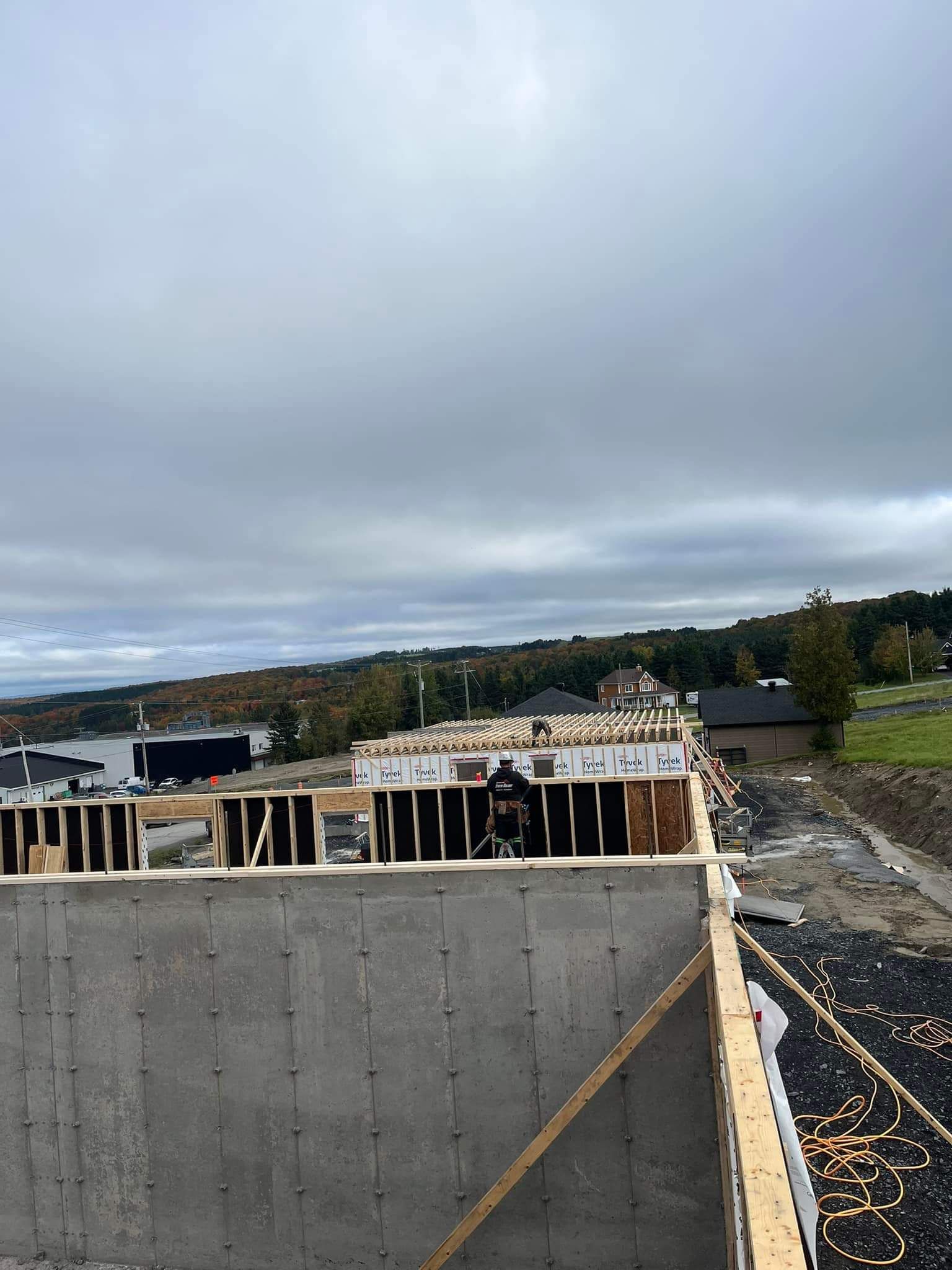 Chantier de construction ; charpente sur fondations en béton. Un homme travaille, ciel nuageux, maisons au loin.