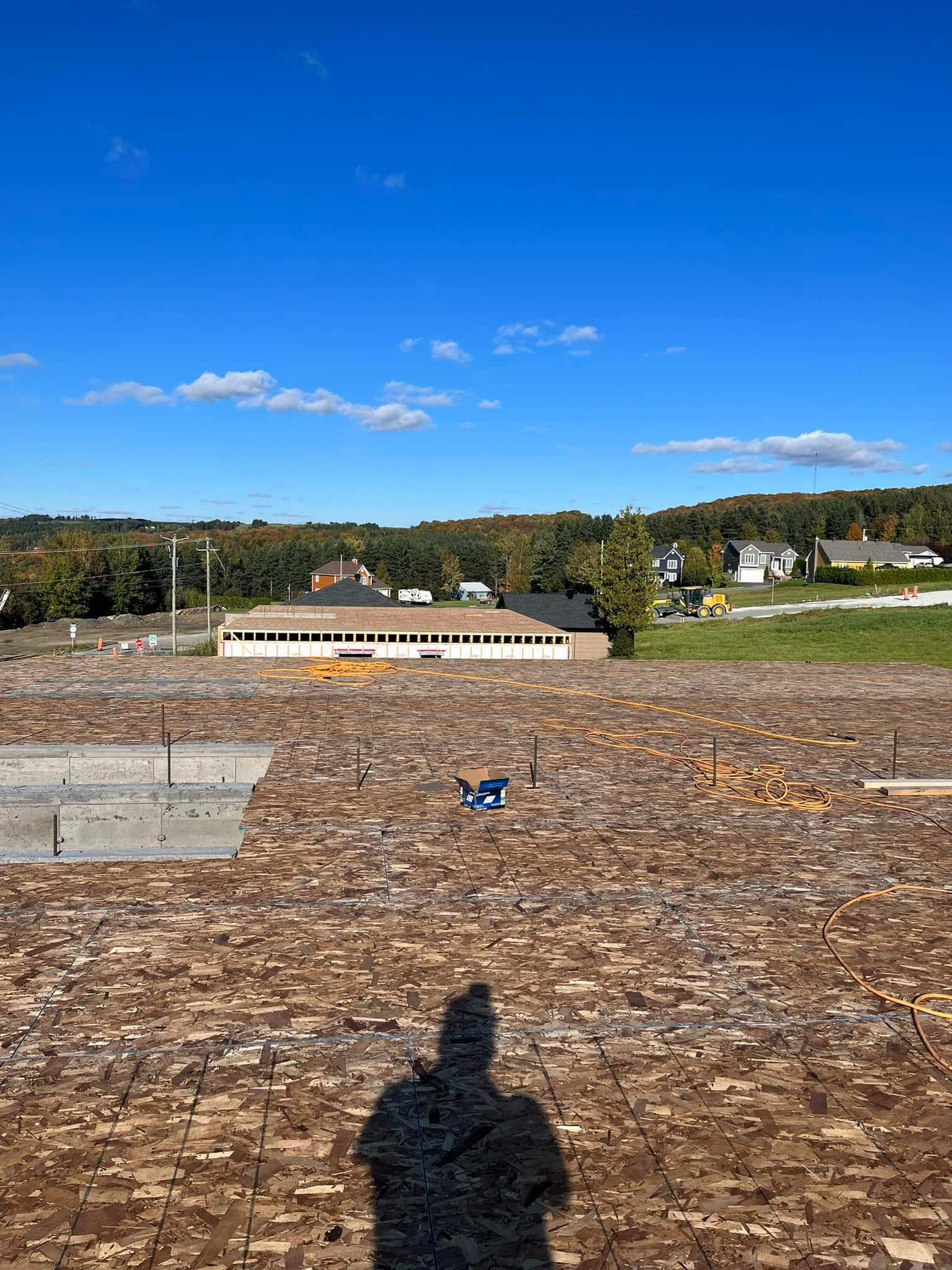 Vue depuis un toit : surface brune, bâtiment au loin, arbres et ciel bleu nuageux.