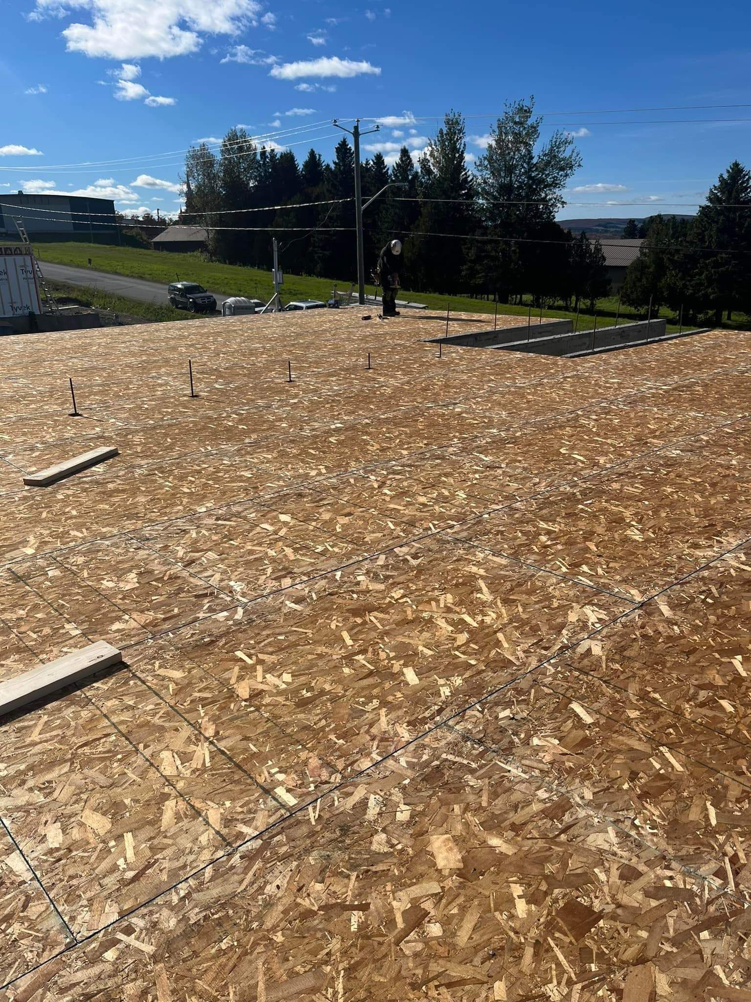 Toiture en panneaux OSB avec une personne debout, ciel bleu dégagé et arbres en arrière-plan.