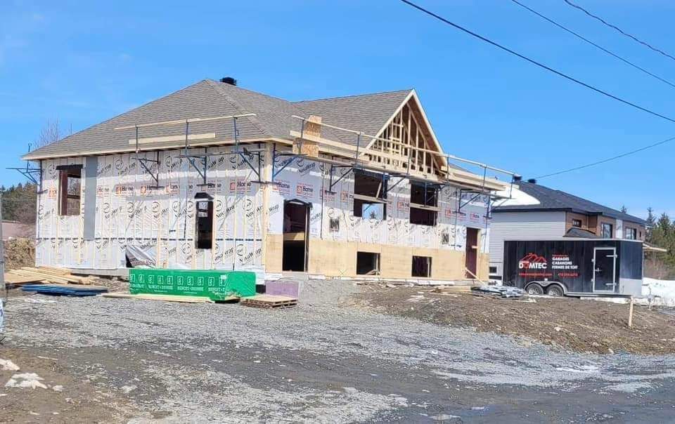 Maison en construction : bâche, revêtement et charpente du toit posés. Matériaux verts et remorque dans la cour.