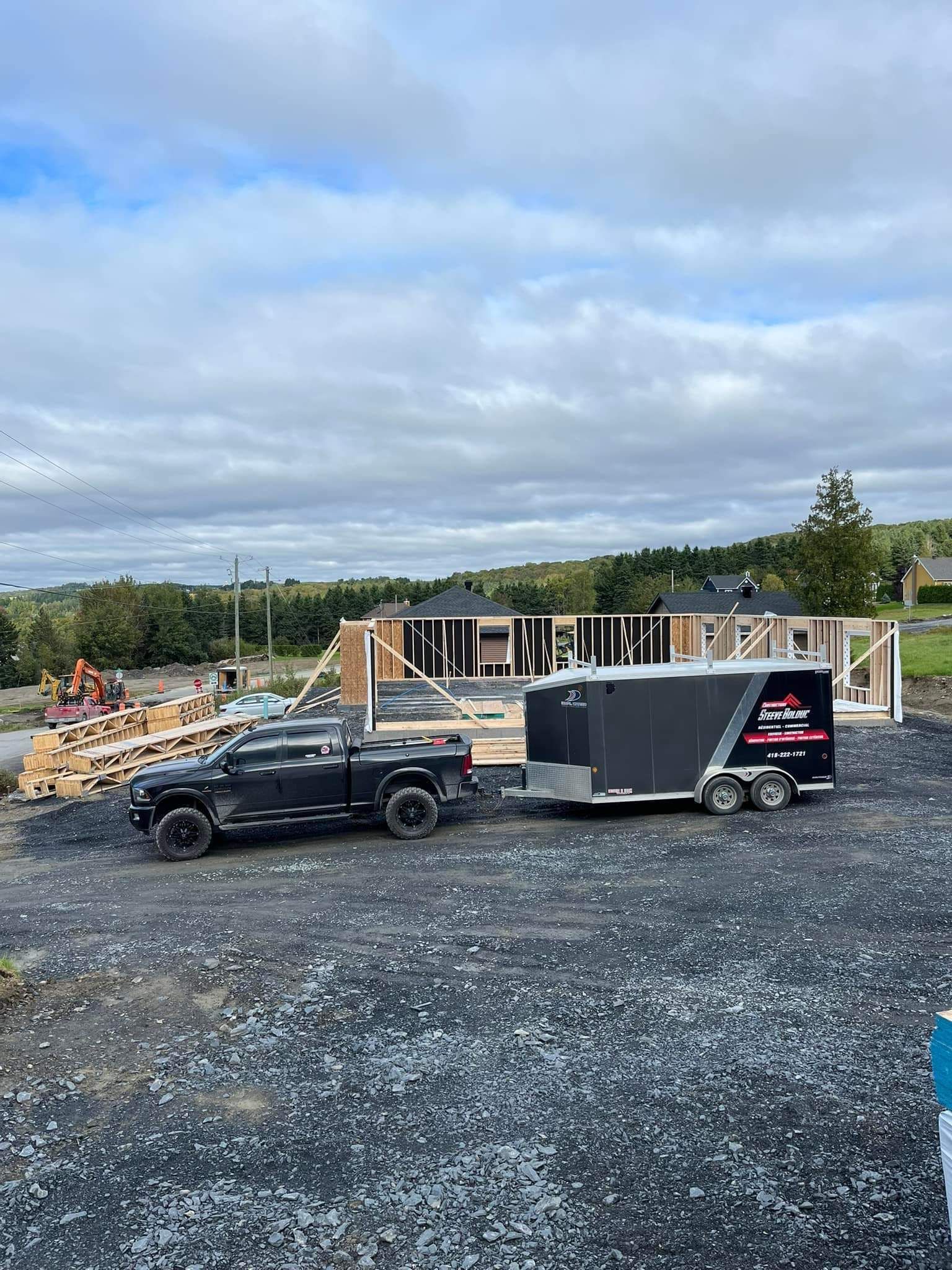 Un camion noir tracte une remorque noire sur un chantier où une maison à ossature partiellement construite. Ciel nuageux.