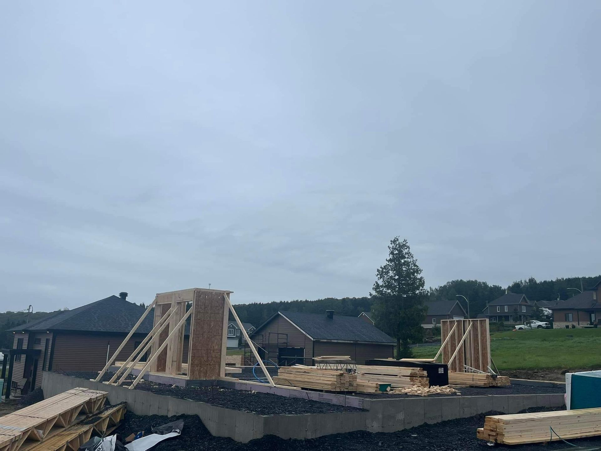 Chantier de construction, charpente en bois d'un bâtiment sur fondations en béton, sous un ciel nuageux.