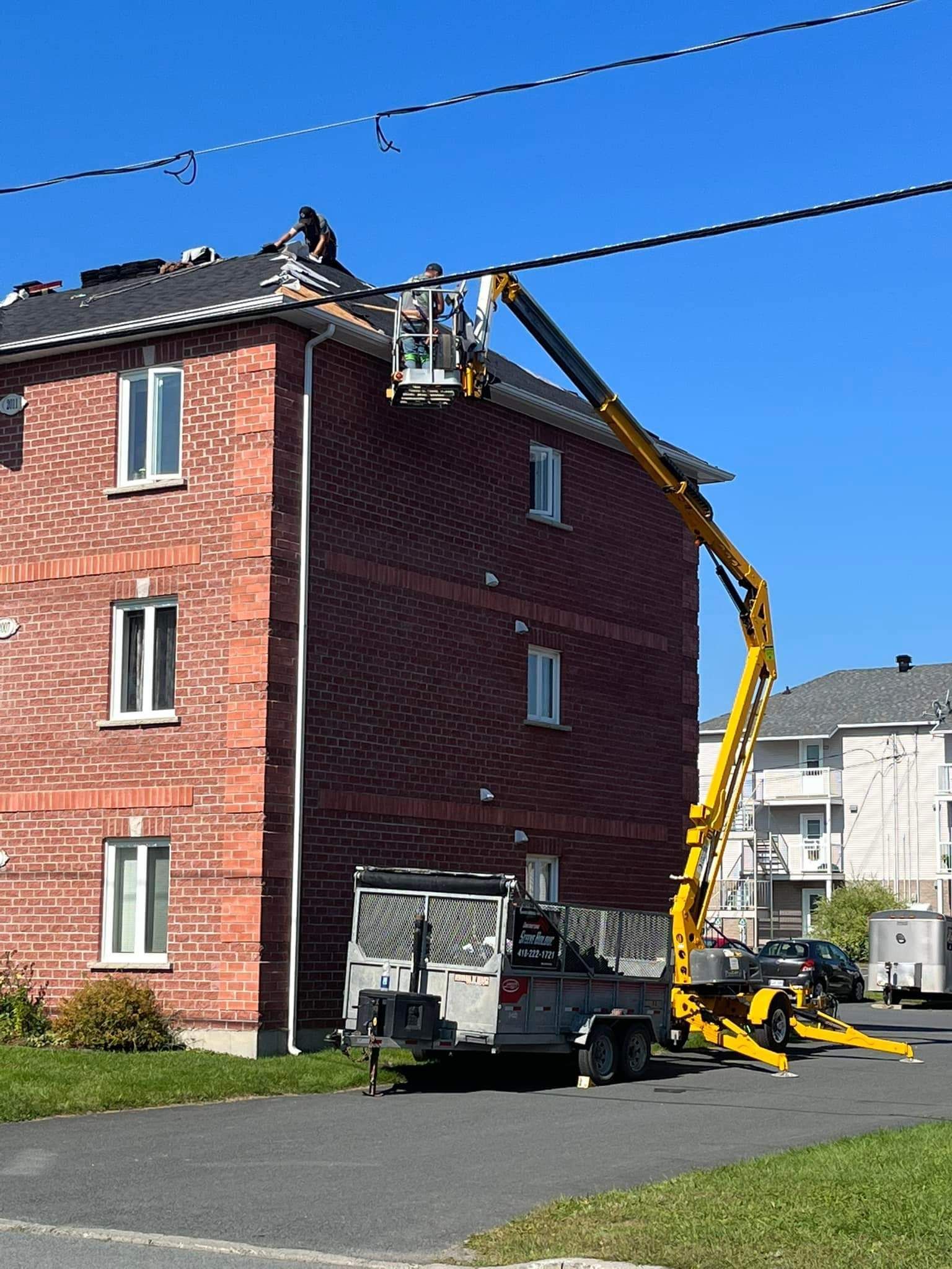 Une nacelle élévatrice répare des câbles près d'un bâtiment en briques, une remorque étant garée en contrebas, sous un ciel dégagé.