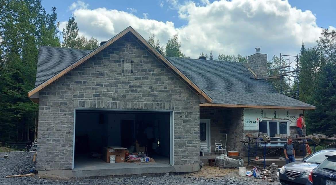 Maison en pierre avec garage ouvert et ouvriers à l'extérieur, entourée d'arbres.