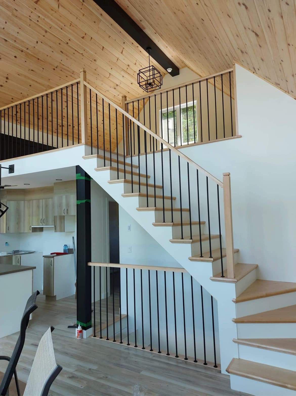 Vue intérieure : Escalier en bois avec rampes verticales noires, menant à une mezzanine. Plafond et murs en bois clair.