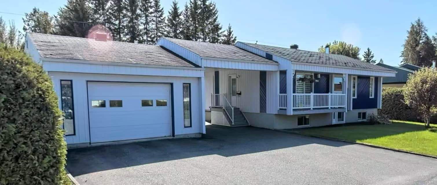 Maison blanche avec garage et allée par une journée ensoleillée. Herbe verte et arbres en arrière-plan.