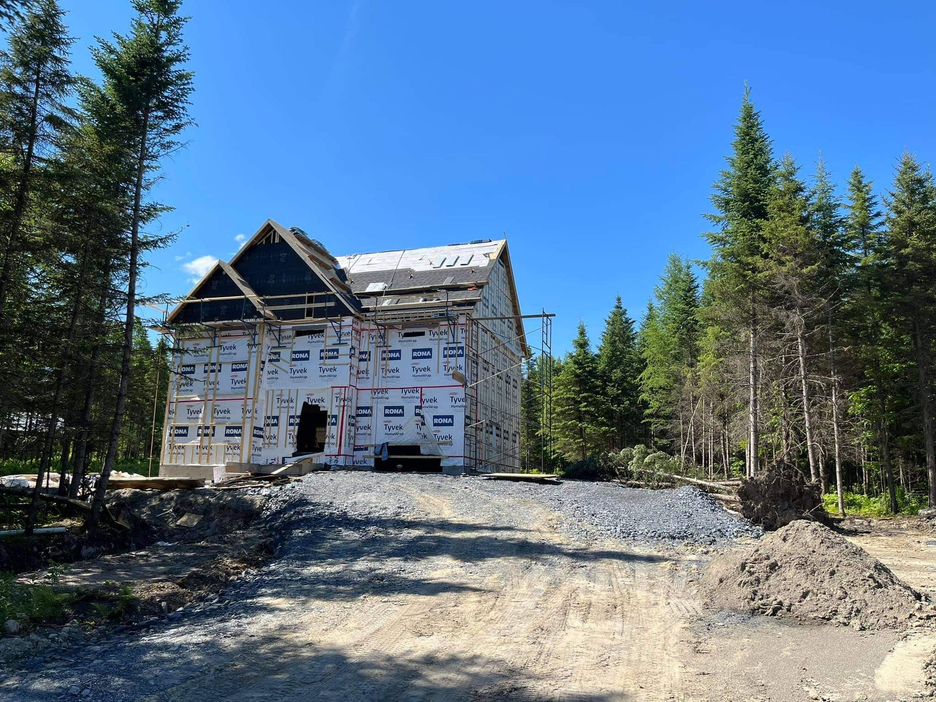 Maison en construction, recouverte d'une bâche blanche, entourée d'arbres et d'une allée de gravier par une journée ensoleillée.