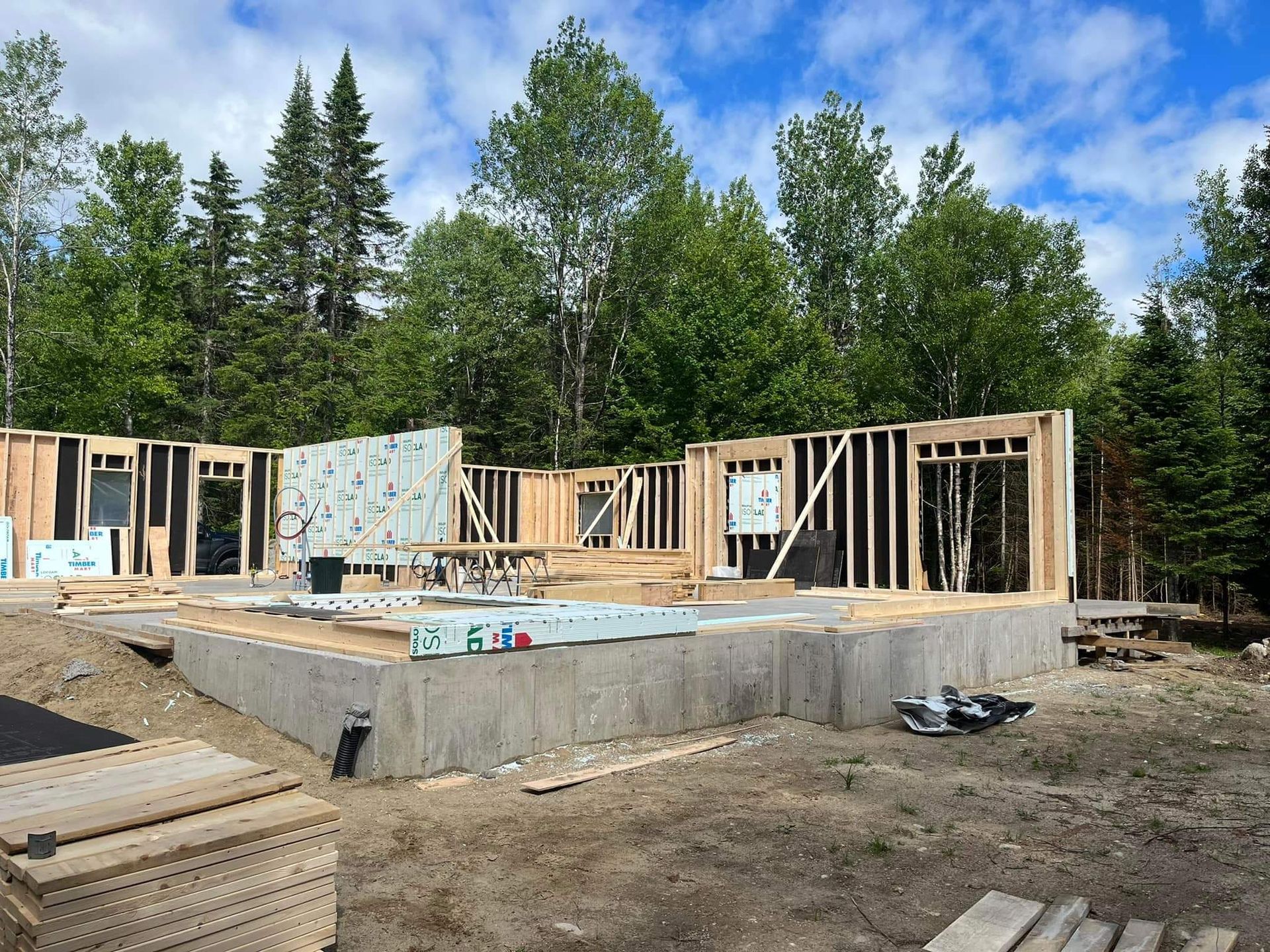 Construction d'une maison : ossature bois sur fondations en béton, avec en arrière-plan des arbres et un ciel bleu.