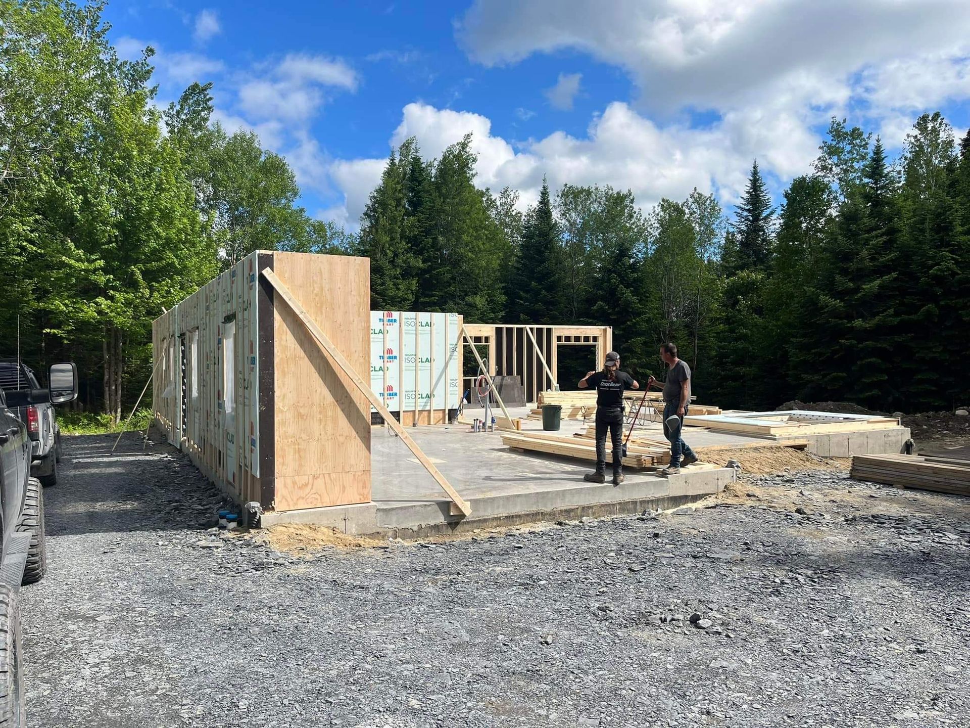 Chantier : deux personnes se tiennent près de la charpente d’un bâtiment. Les murs et les fondations sont visibles.
