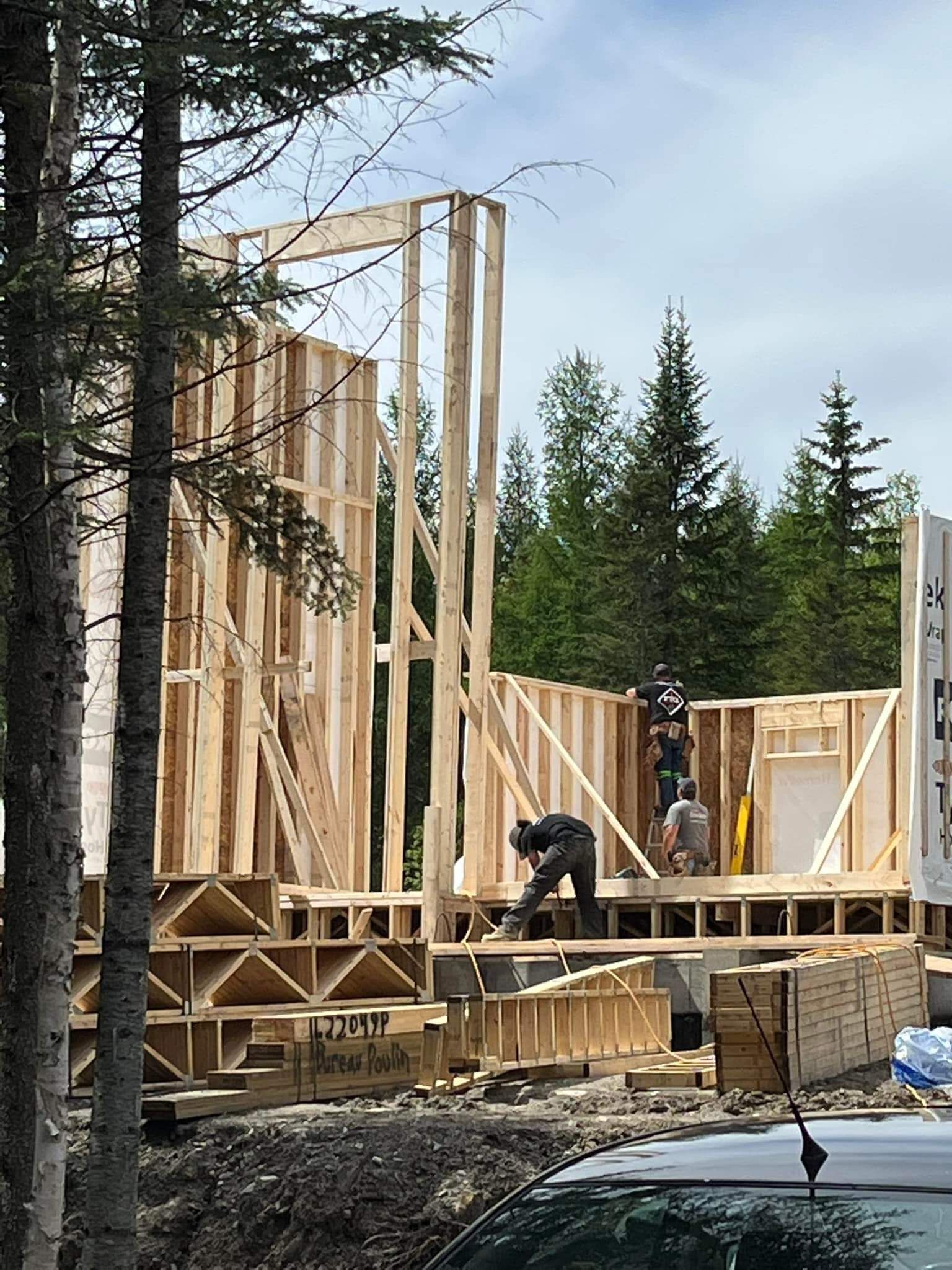 Construction d'un bâtiment à ossature bois ; des ouvriers construisent les murs sur des fondations, avec des arbres en arrière-plan.