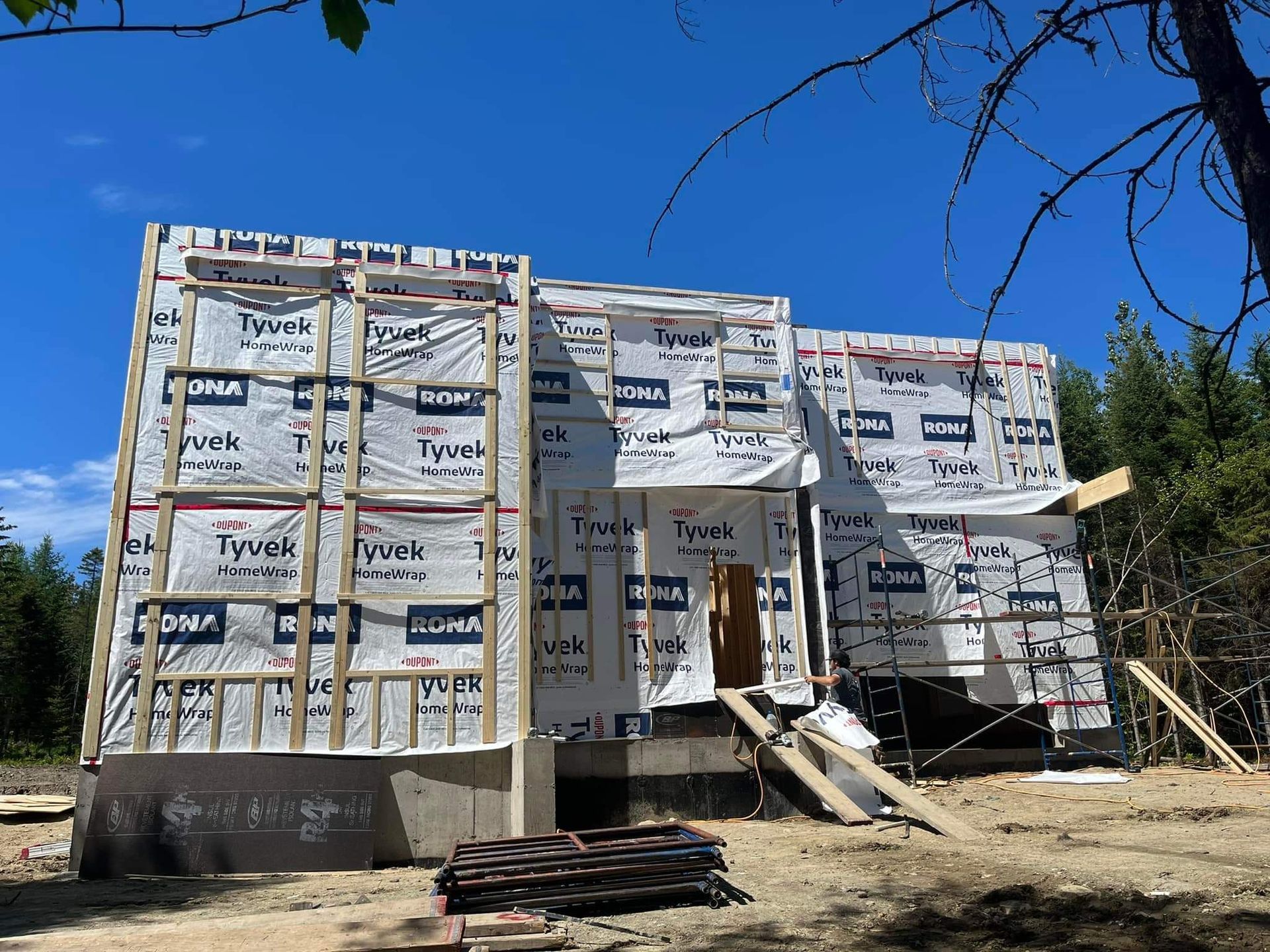 Maison en construction avec bâche Tyvek, ossature bois et fondations en béton ; à l'extérieur par une journée ensoleillée.