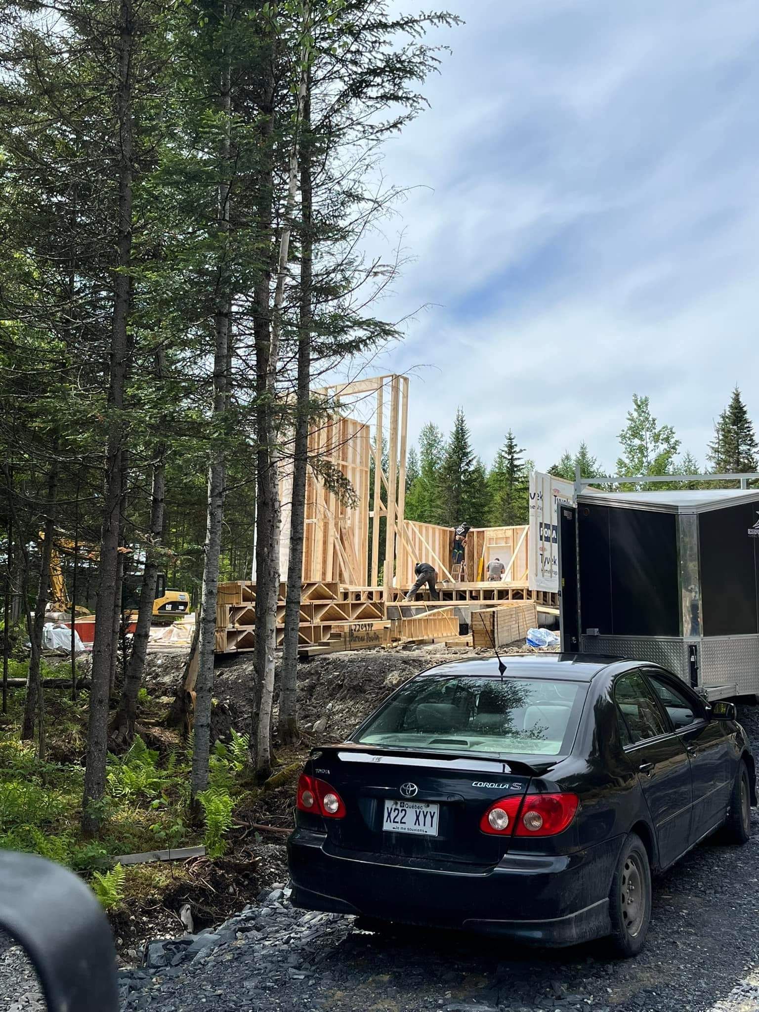 Voiture garée près d'une maison en bois en construction dans une zone boisée. Le ciel est couvert.